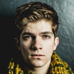 Close-up portrait of a young adult male with a serious expression against a dark textured wall.